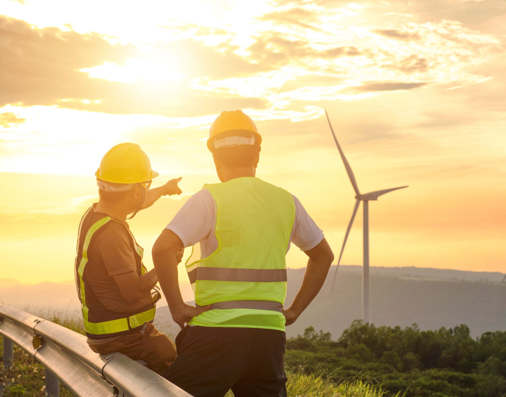 Two men in safety vests and helmets inspect wind turbines outdoors.