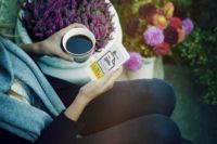 Person hält eine Tasse Kaffee und ein Smartphone, umgeben von Blumen im Freien.